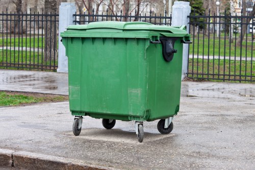 A large green plastic wheeled rubbish bin with a black handle and a closed lid, situated on a damp asphalt surface in an outdoor urban environment. The bin is positioned in front of a black metal fence with vertical bars, behind which there is a paved walkway, some grass patches, and trees with bare branches, indicating a park or residential area in Shepherds Bush. The scene is illuminated by overcast natural light, and the surface around the bin appears slightly wet, suggesting recent rain. This image is relevant for a rubbish removal service such as Business Waste Removal Shepherds Bush, highlighting the type of waste container typically handled in local waste management operations.
