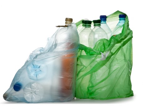 A collection of various used plastic bottles, including clear, green, and blue-tinted containers, loosely placed inside a partially open green plastic rubbish bag. The bottles are of different shapes and sizes, with some cap closures visible, and the plastic surfaces exhibiting a glossy, slightly crinkled texture. The scene appears to be set against a plain white background, emphasizing the bottles and the bag, which are positioned in the foreground. This arrangement suggests a typical scene associated with waste collection or recycling, relevant to rubbish removal services, particularly in the Shepherds Bush area. The image illustrates the common practice of collecting plastic waste for disposal or recycling, aligned with services provided by companies like Business Waste Removal Shepherds Bush, operating within the local community postcode and surroundings.